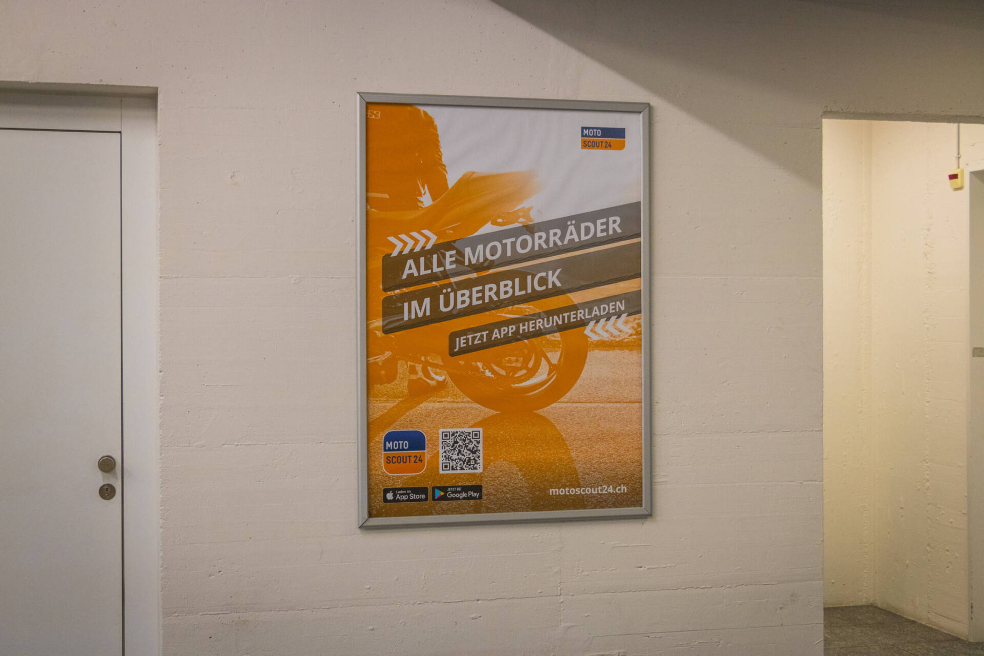 An einer hellen Wand hängt ein orange gestaltetes Plakat mit der Aufschrift «Alle Motorräder im Überblick – jetzt App herunterladen», dazu ein QR-Code, App-Store-Badges und die Adresse motoscout24.ch.