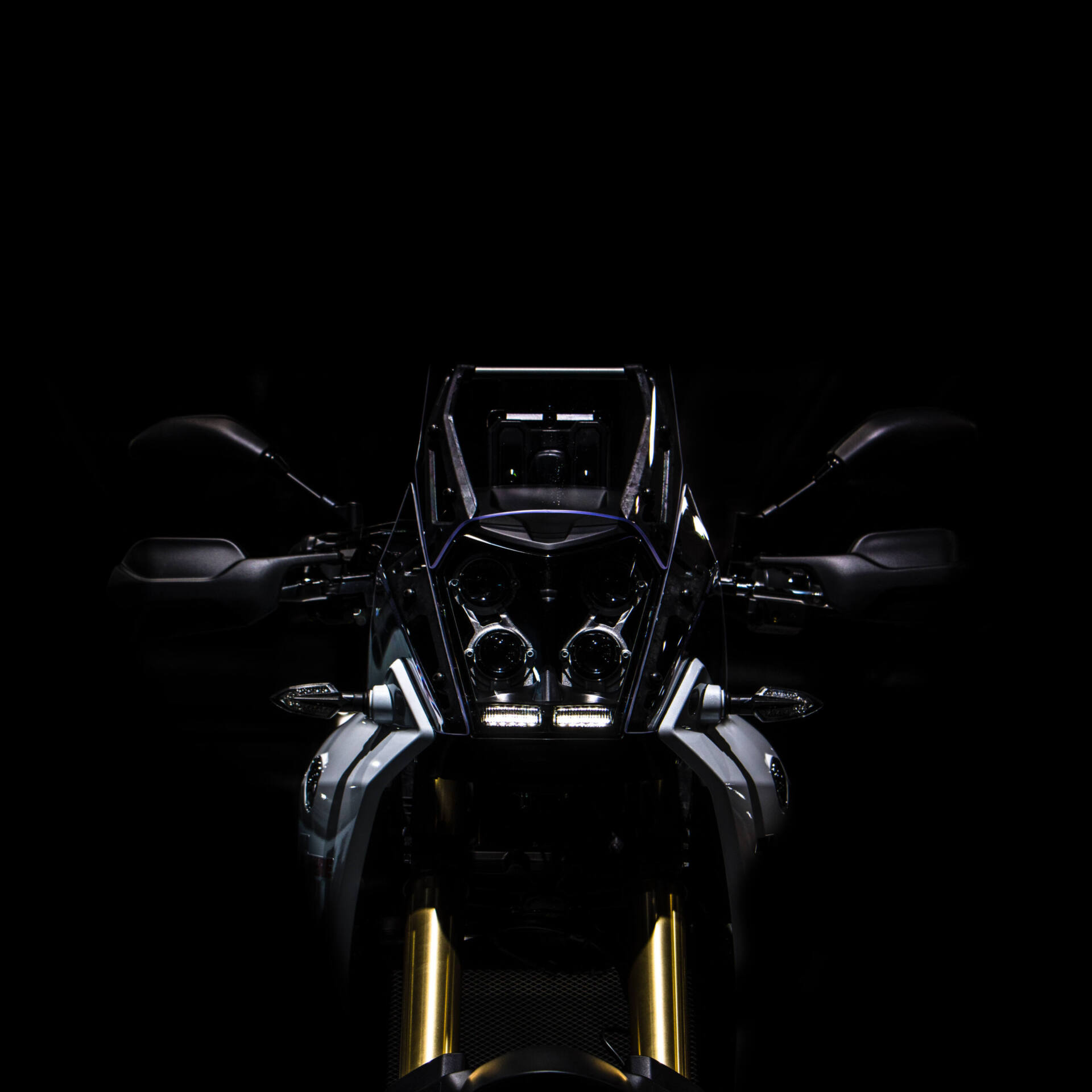 Frontmaske einer Yamaha Ténéré im Dunkeln. Vier runde Scheinwerfer und eine schmale LED-Leiste über der goldenen Gabel wirken wie ein Gesicht mit mehreren Augen.