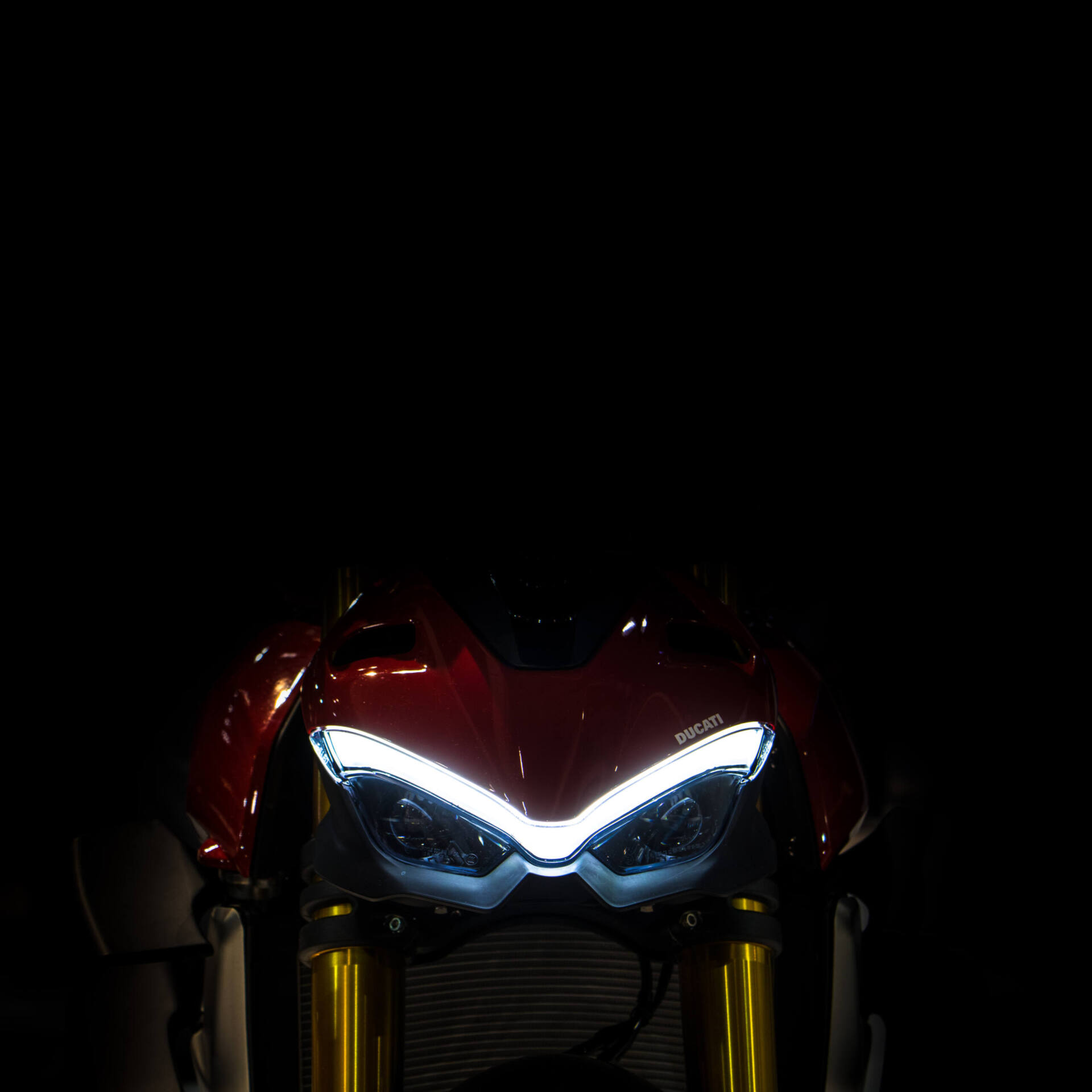 Front einer roten Ducati Streetfighter im Dunkeln. Das V-förmige LED-Tagfahrlicht über der goldenen Gabel wirkt wie ein Paar zusammengezogener, finsterer Augenbrauen.