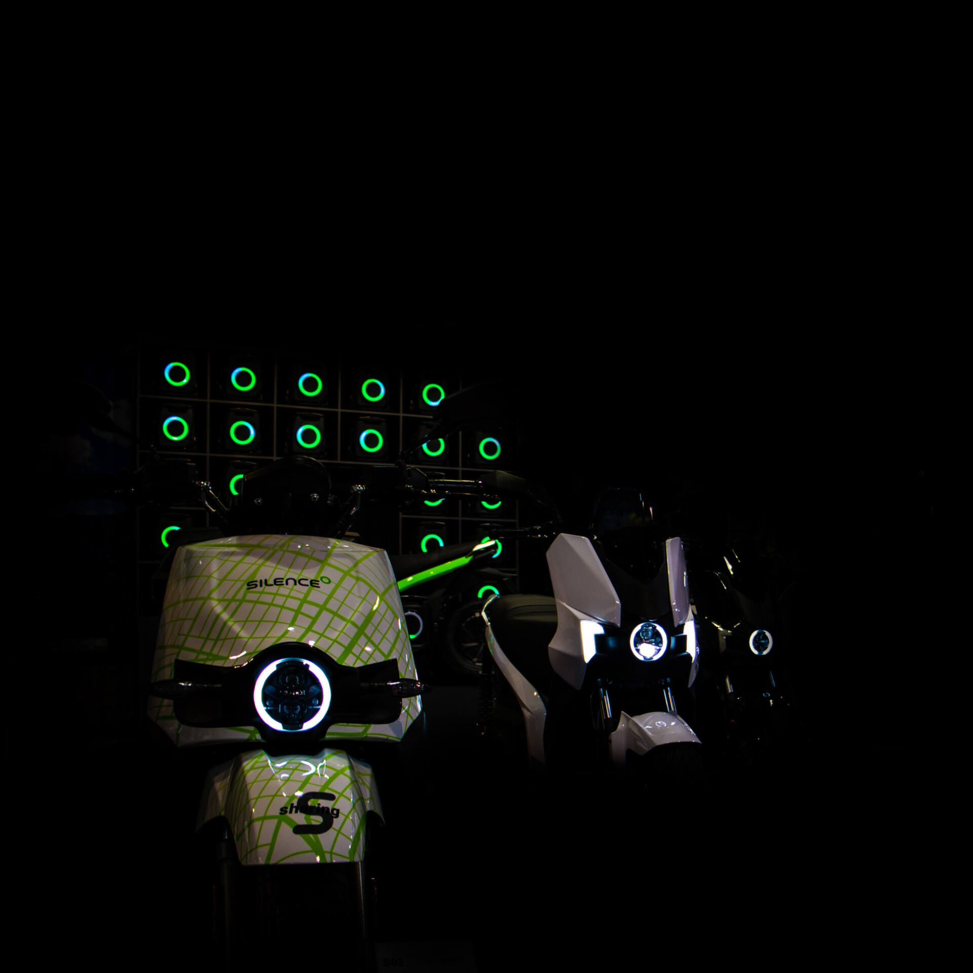 Weiss-grüne Silence-Elektroroller an einem dunklen Messestand. Der runde LED-Scheinwerfer des vordersten Rollers leuchtet wie ein einzelnes Auge vor einer Wand aus grünen Lichtringen.