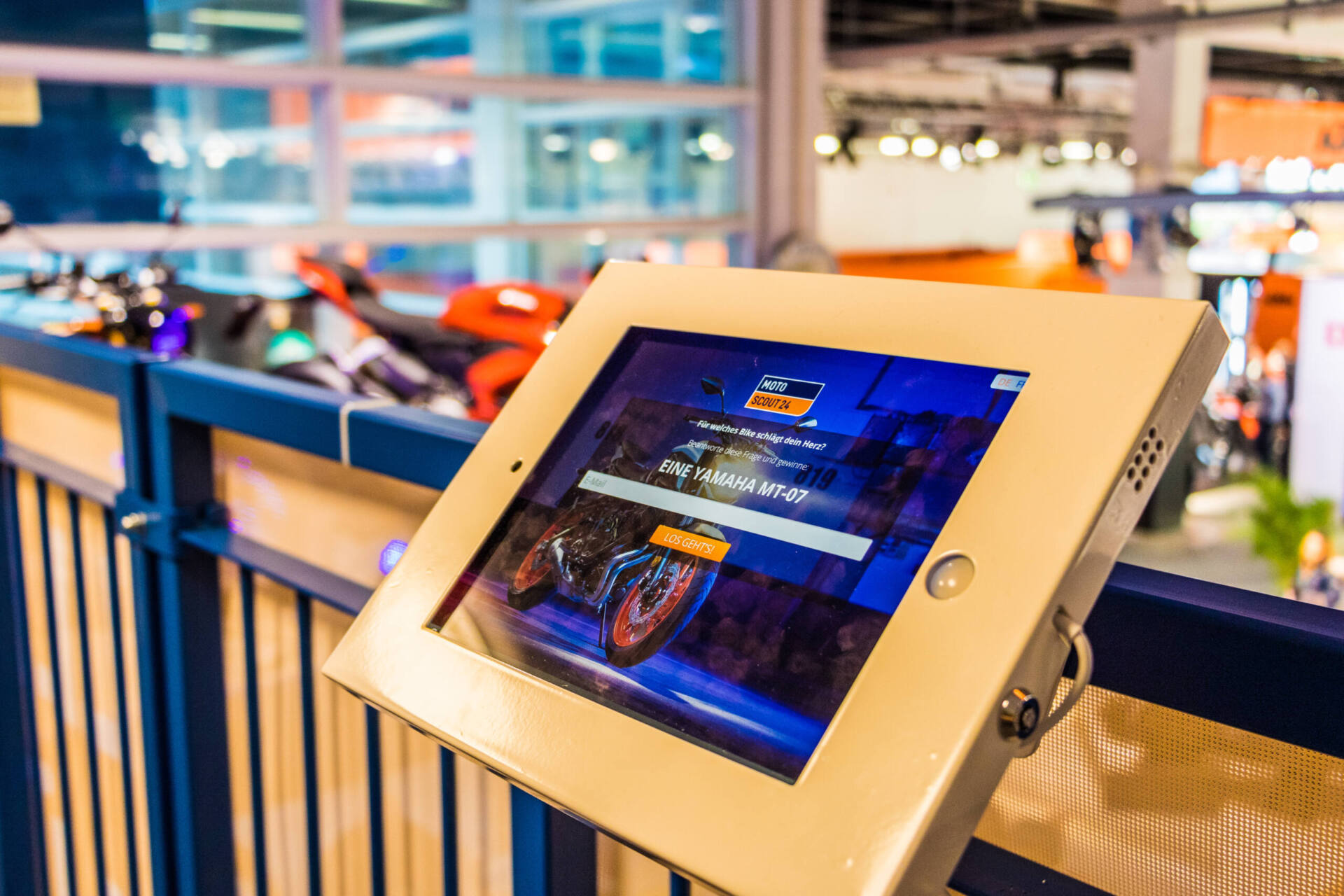 Ein in einer beigen Halterung montiertes iPad am Messegeländer zeigt den MotoScout24-Wettbewerb mit dem Hinweis «Eine Yamaha MT-07». Im unscharfen Hintergrund stehen Motorräder in der Messehalle.