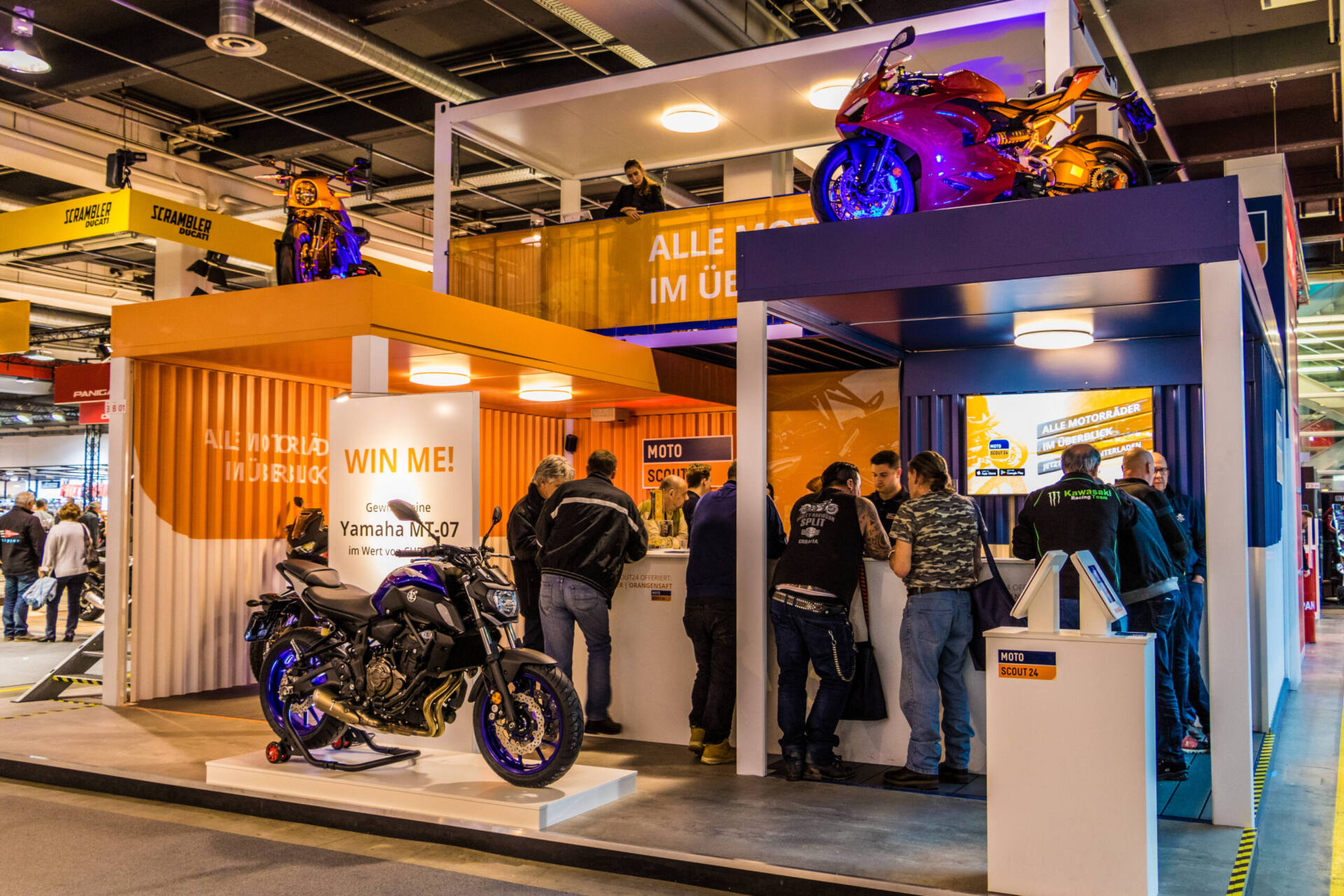 Der MotoScout24-Messestand in oranger Container-Optik mit Besuchern am Stand. Auf einem Podest steht ein blaues Yamaha MT-07 mit der Aufschrift «Win me!», auf dem oberen Deck eine rote Ducati.