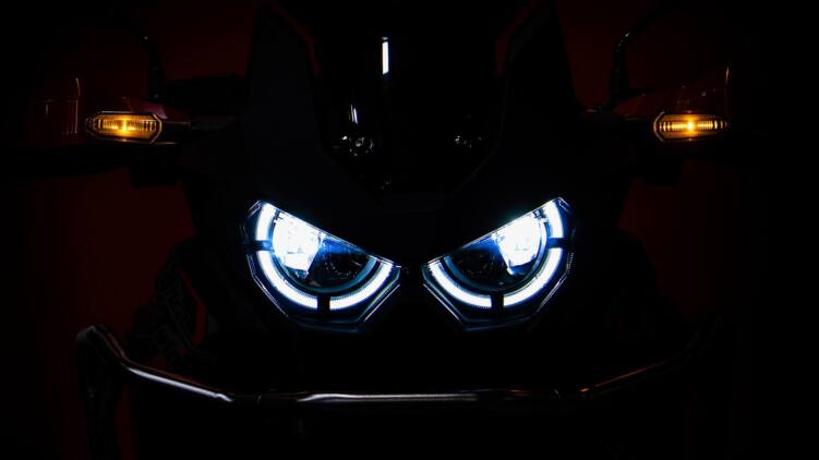 Frontale Nahaufnahme eines Motorrads im Dunkeln. Zwei hell blau-weiss leuchtende LED-Scheinwerfer und die bernsteinfarbenen Blinker lassen die Front wie ein Gesicht wirken – fremd, fast insektenartig.