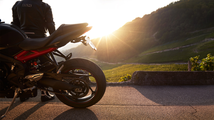 Motorradfahrer in Lederkombi von hinten vor seiner Triumph Street Triple R 675, Blick in den Sonnenuntergang über den Weinbergen am Bielersee.