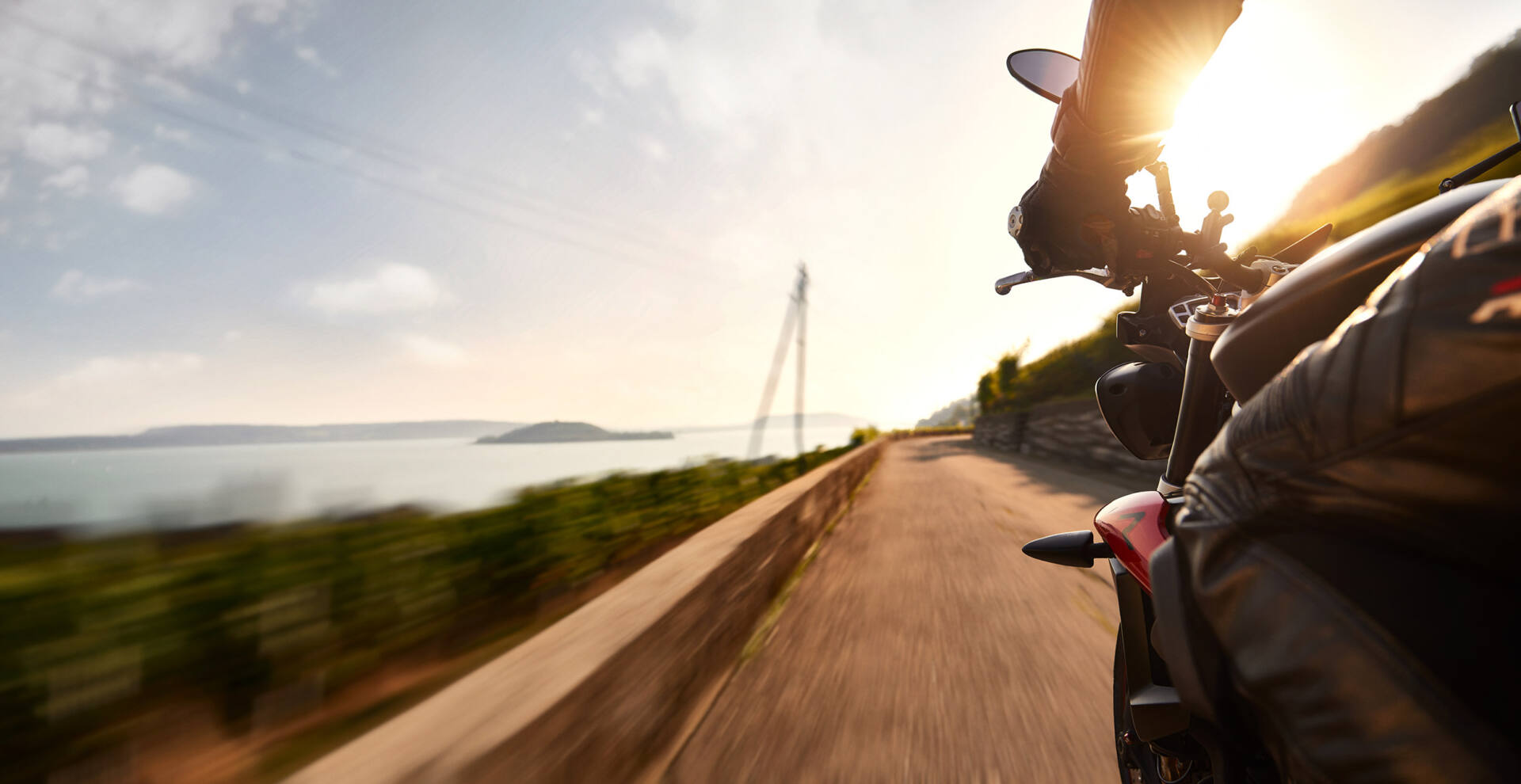 Motorrad in Fahrt auf einer Strasse hoch über dem Bielersee, Abendsonne im Gegenlicht, Weinberge am Hang und Wasser mit Insel im Hintergrund.