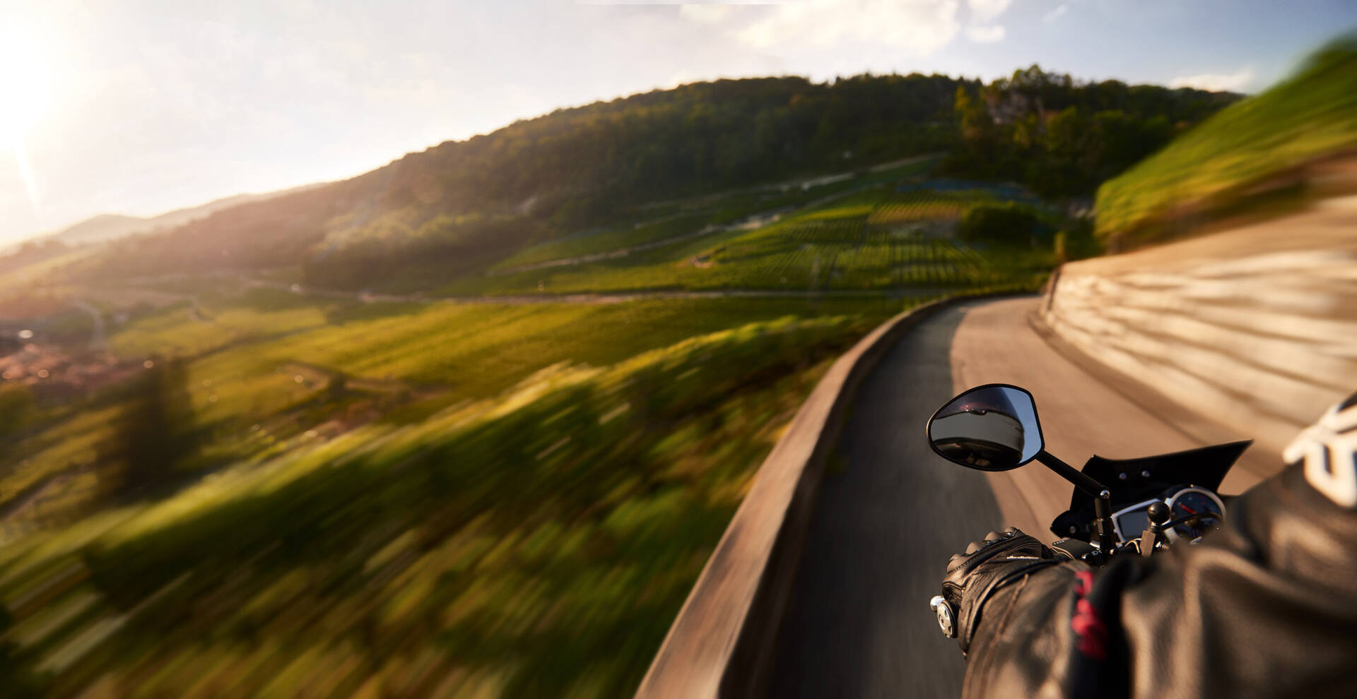 Blick über Lenker und Spiegel eines fahrenden Motorrads in eine Kurve, Rebhänge im warmen Abendlicht, seitlich verwischt durch die Bewegung.