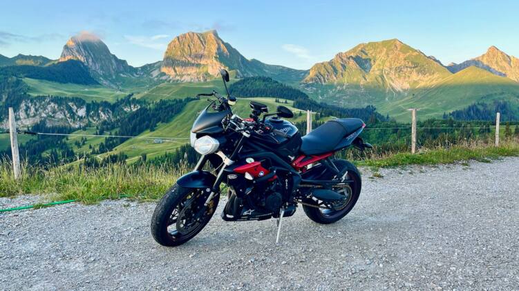 Schwarze Triumph Street Triple R 675 mit roten Akzenten auf dem Seitenständer am Gurnigelpass, dahinter die vom Morgenlicht beschienenen Gipfel der Gantrisch-Kette über grünen Alpweiden.