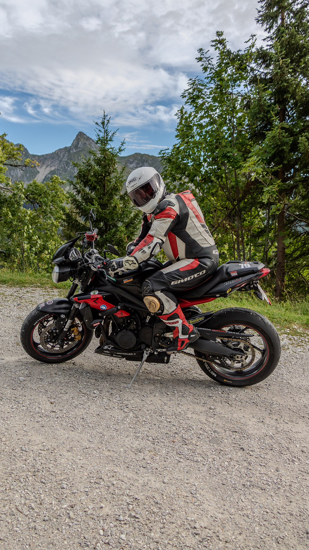 Ein Motorradfahrer in rot-weiss-schwarzer Lederkombi und weissem Helm sitzt auf einem schwarz-roten Naked Bike auf einem Kiesplatz, darüber viel Himmel, dahinter Nadelwald und ein Berggipfel.