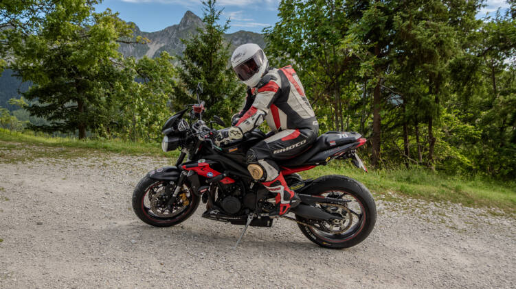 Ein Motorradfahrer in rot-weiss-schwarzer Lederkombi und weissem Helm sitzt auf einem schwarz-roten Naked Bike auf einem Kiesplatz, dahinter Nadelwald und ein Bergpanorama.