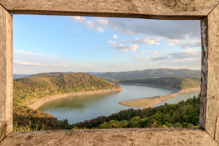 Blick vom Elsterberg durch einen verwitterten Holzrahmen auf den Edersee in Richtung Staumauer, im warmen Licht. Der See windet sich zwischen bewaldeten Hügeln, entlang der Ufer ziehen sich breite helle Streifen freigelegten Bodens, rechts ragt eine lange Sandbank ins Wasser. Darüber blauer Himmel mit Wolken.