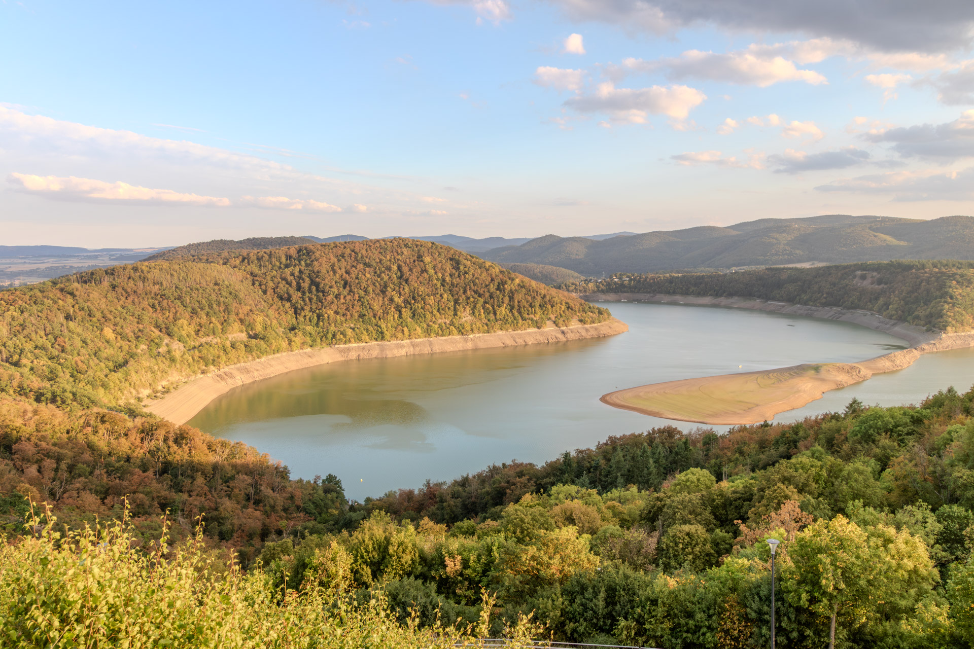 Blick vom Elsterberg auf den Edersee in Richtung Staumauer. Der See liegt in einer weiten Schleife zwischen bewaldeten Hügeln, an den Ufern breite helle Streifen freigelegten Bodens, rechts eine lange Sandbank im Wasser. Im Vordergrund Büsche und eine Strassenlaterne, darüber blauer Himmel mit Wolken im warmen Licht.