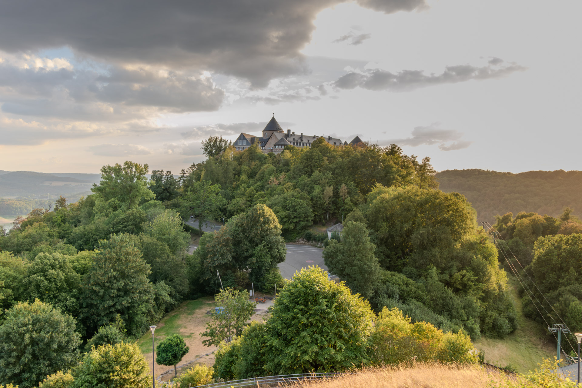 Schloss Waldeck mit seinem runden Turm auf einem dicht bewaldeten Hügel, darüber dunkle Wolken und heller Abendhimmel. Unten zwischen den Bäumen ein Parkplatz und eine Wiese, rechts ein Mast mit den Seilen der Seilbahn.