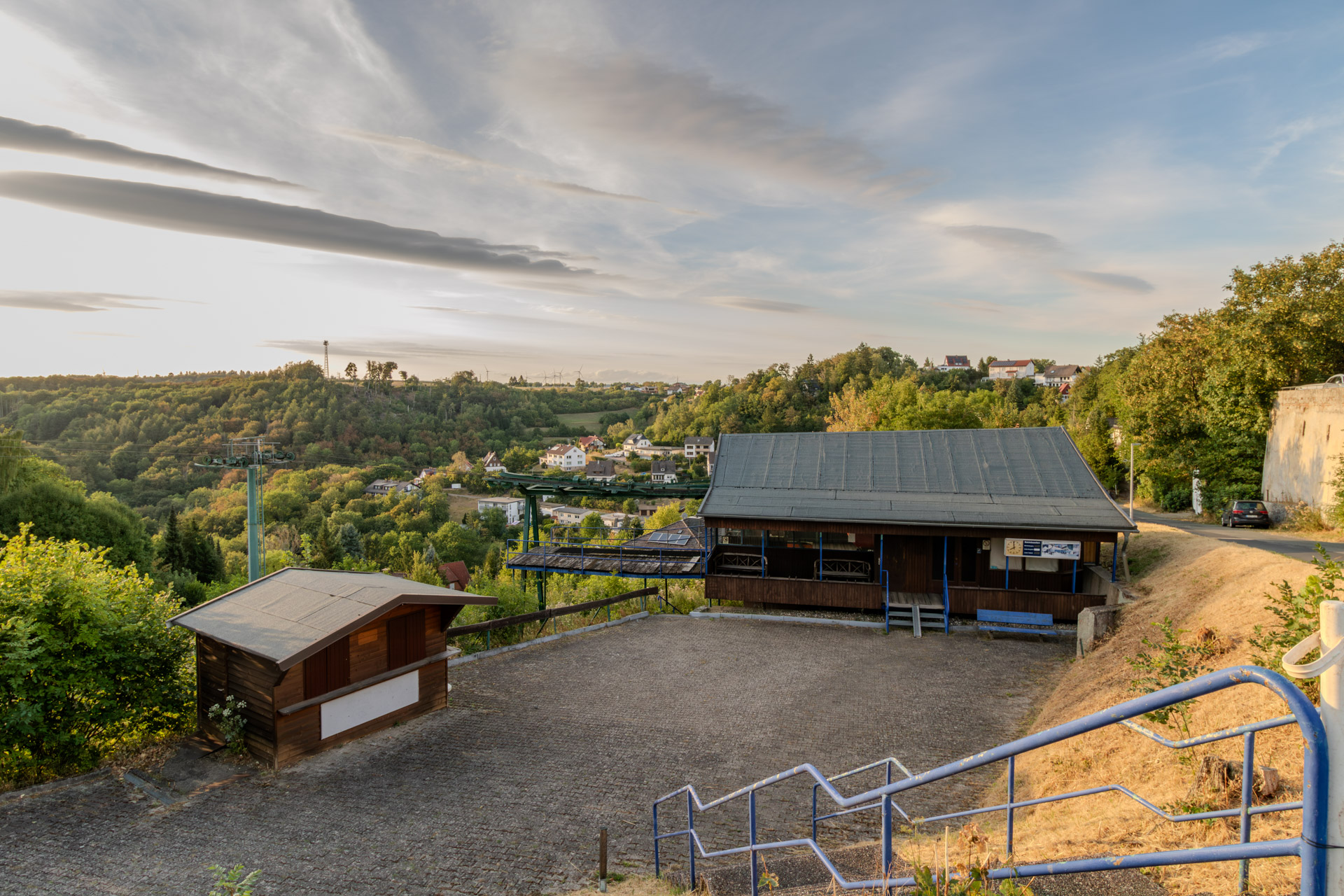 Die Bergstation der Seilbahn, ein dunkles Holzgebäude mit flachem Satteldach, davor ein leerer gepflasterter Platz und ein geschlossener Kiosk aus Holz. Rechts eine Treppe mit blauem Geländer und vertrocknetes Gras, links die grünen Stützen der Seilbahn, die ins Tal führen. Dahinter Häuser des Ortes zwischen bewaldeten Hügeln.