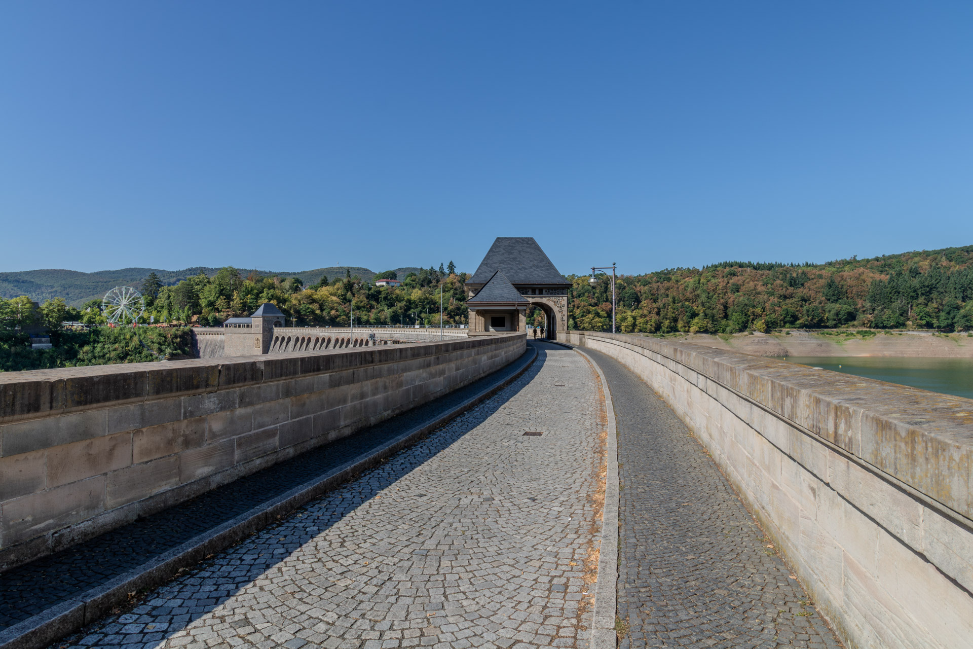 Gepflasterter Weg auf der Krone der Staumauer zwischen zwei Brüstungen aus Sandstein, in der Mitte ein Turm mit spitzem Schieferdach und Durchgangsbogen, durch den zwei Personen gehen. Links der geschwungene Verlauf der Staumauer mit ihren Bögen und in der Ferne ein Riesenrad am Sperrmauervorplatz in Hemfurth-Edersee, rechts der Edersee mit breitem freigelegtem Ufer. Wolkenloser blauer Himmel.