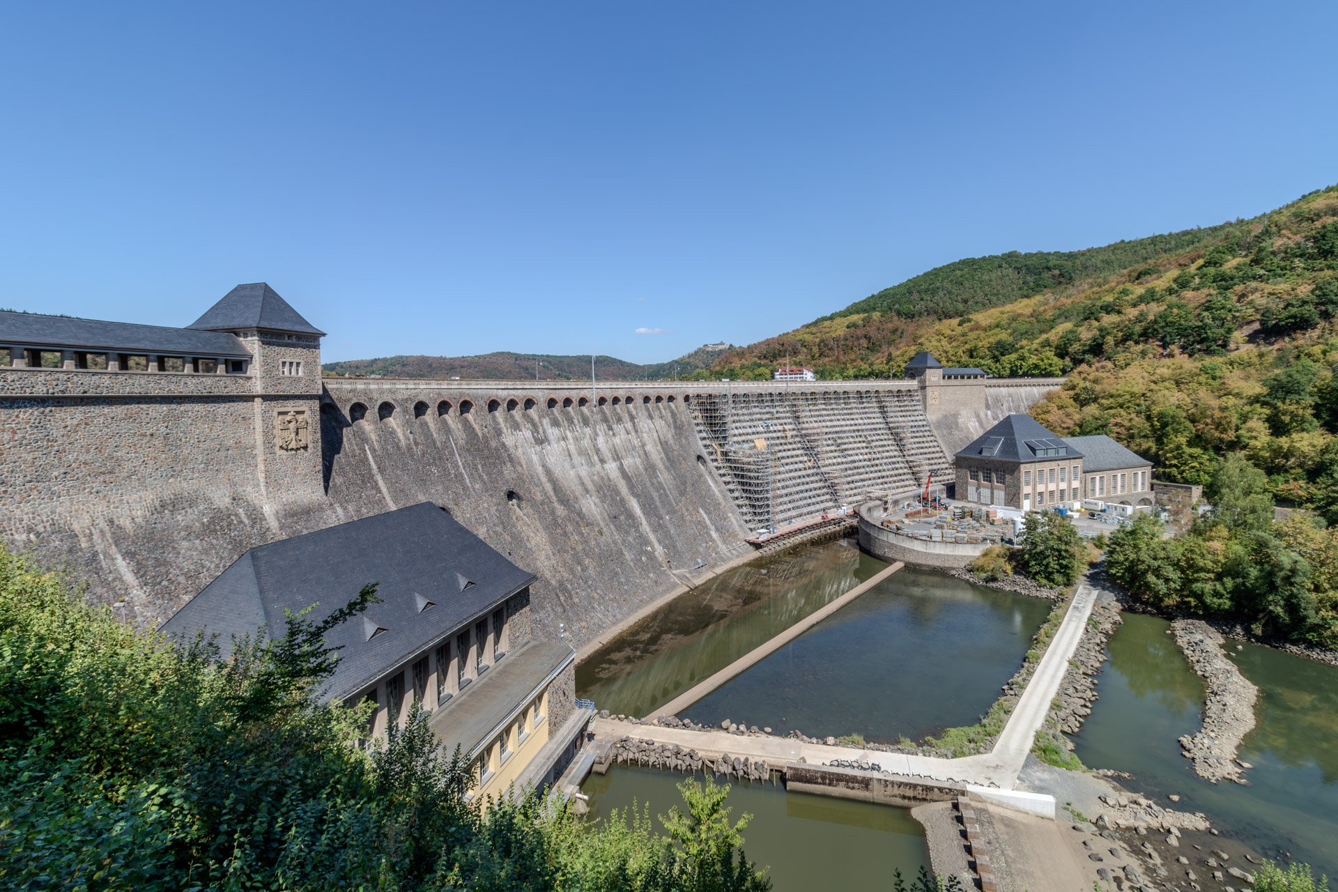 Die geschwungene Staumauer der Edertalsperre von der Talseite aus, oben eine Reihe von Bögen, an beiden Enden Türme mit Schieferdach. Der rechte Teil der Mauer ist komplett eingerüstet, davor eine Baustelle mit Material und Baumaschinen. Im Vordergrund Kraftwerksgebäude und ein Becken mit wenig Wasser, in der Ferne auf einem Hügel Schloss Waldeck.