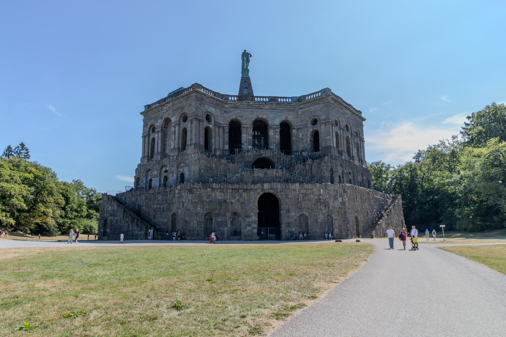 Das Oktogon im Bergpark Wilhelmshöhe von der Rückseite, ein mächtiger Bau aus dunklem Stein mit Rundbögen und seitlichen Treppen, obenauf die Pyramide mit der Herkules-Statue. Davor eine Wiese und ein breiter Weg, auf dem einige Besucher unterwegs sind, links und rechts Bäume unter blauem Himmel.