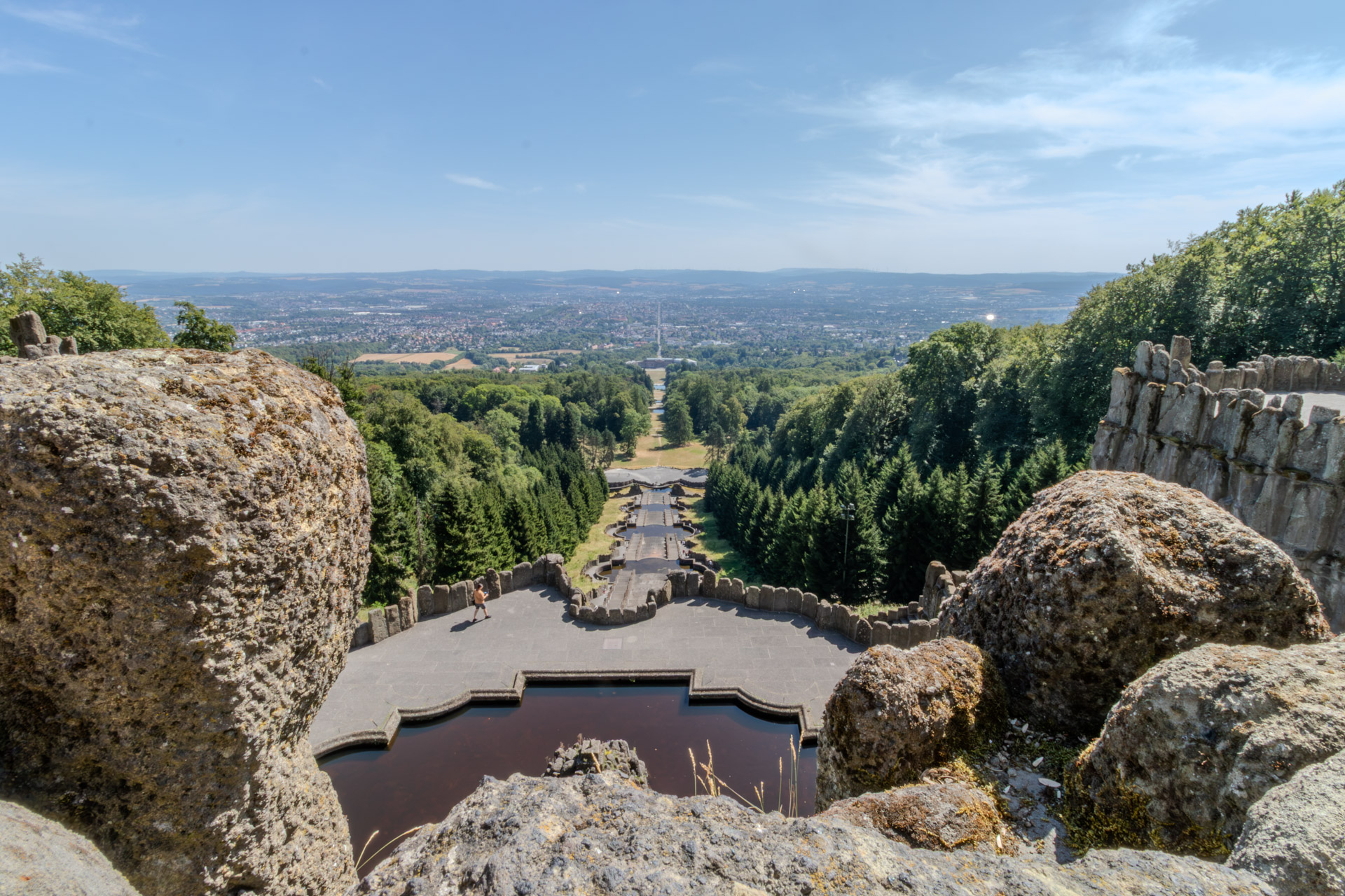 Blick vom Herkules zwischen grossen Felsbrocken hinunter über die Kaskaden, eine lange Achse aus Treppen und Becken, die schnurgerade durch den Wald führt. Am Ende der Achse Schloss Wilhelmshöhe, dahinter breitet sich Kassel bis zum Horizont aus. Im Vordergrund ein Wasserbecken, auf der Plattform davor eine einzelne Person.
