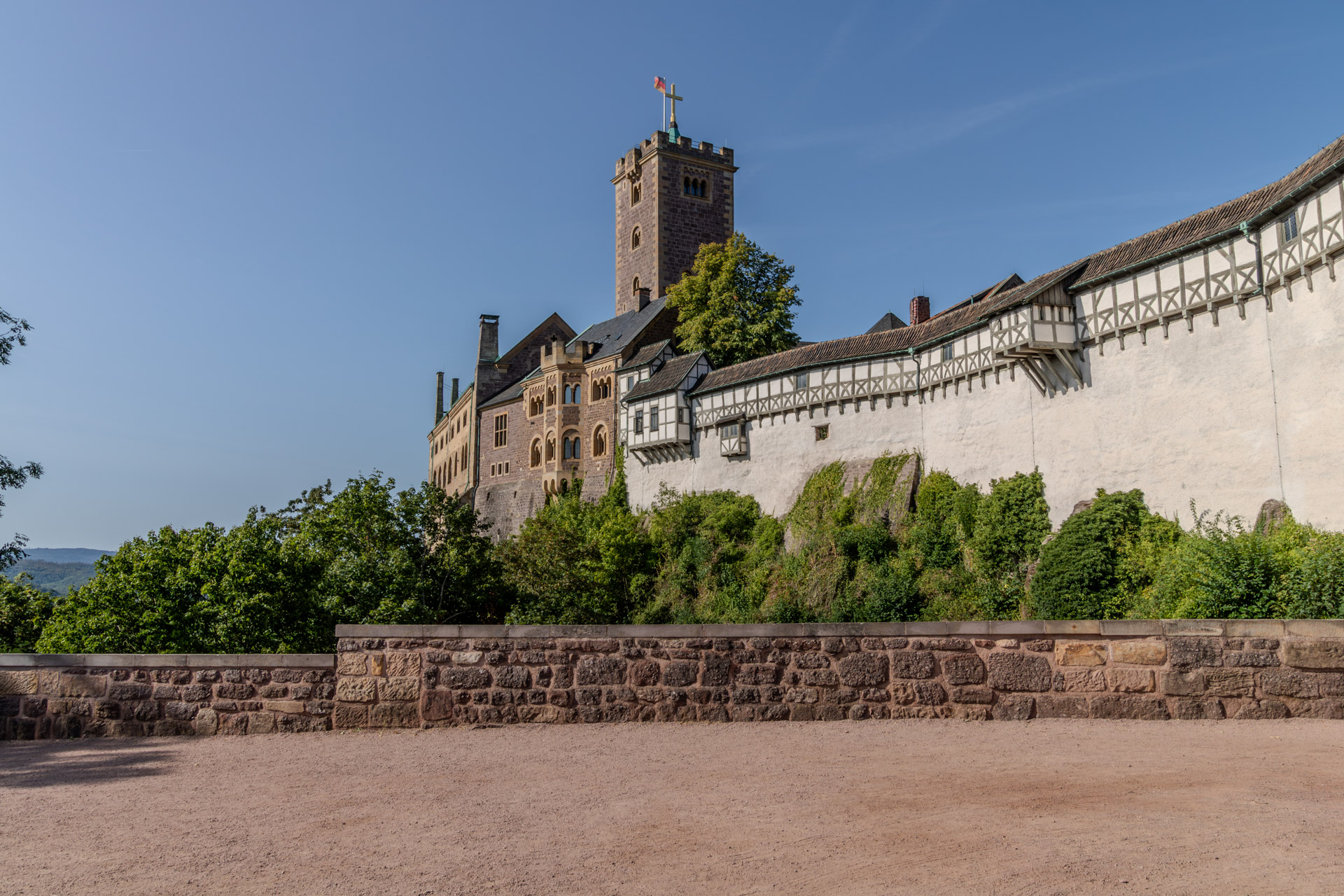 Die Wartburg von unten gesehen, links der quadratische Bergfried mit goldenem Kreuz und Fahne, daneben Gebäude aus Stein mit Rundbogenfenstern. Rechts zieht sich die weisse Burgmauer mit einem Wehrgang aus Fachwerk den Hang entlang. Im Vordergrund ein Kiesplatz und eine niedrige Mauer aus Bruchstein, dahinter dichtes Grün.