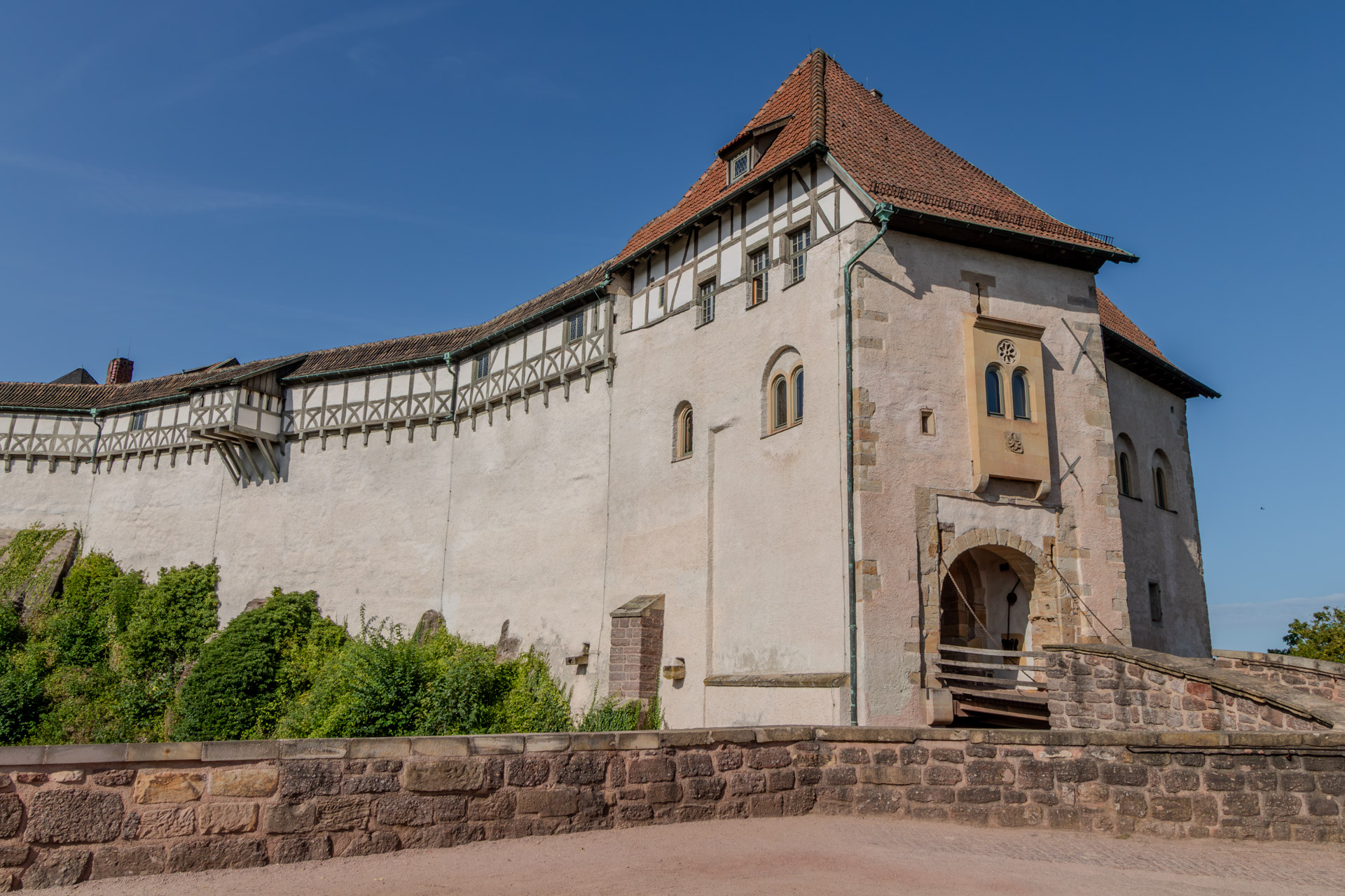 Das Torhaus der Wartburg mit rotem Ziegeldach und Fachwerk im Obergeschoss, unten ein Rundbogentor mit hölzerner Zugbrücke und Ketten. Links schliesst die weisse Burgmauer mit Fachwerk-Wehrgang an, davor Büsche und eine Mauer aus Bruchstein unter tiefblauem Himmel.