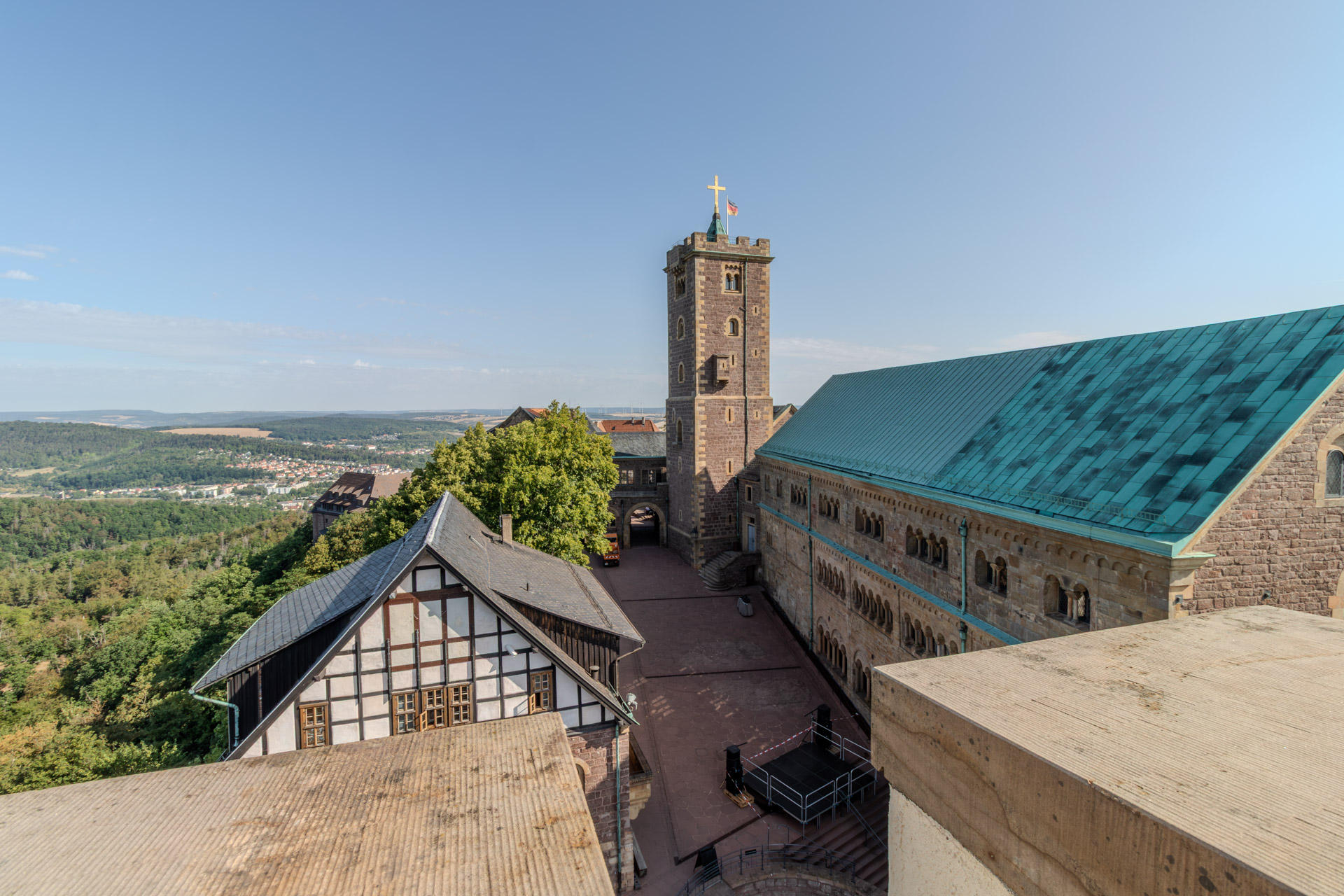 Blick von oben in den Burghof der Wartburg, in der Mitte der Bergfried mit goldenem Kreuz und Deutschlandfahne. Rechts der Palas mit seinen Rundbogenfenstern und dem grünen Kupferdach, links ein Fachwerkhaus mit Schieferdach. Im Hof eine aufgebaute Bühne, links hinten der Blick über bewaldete Hügel bis zu den Häusern von Eisenach.