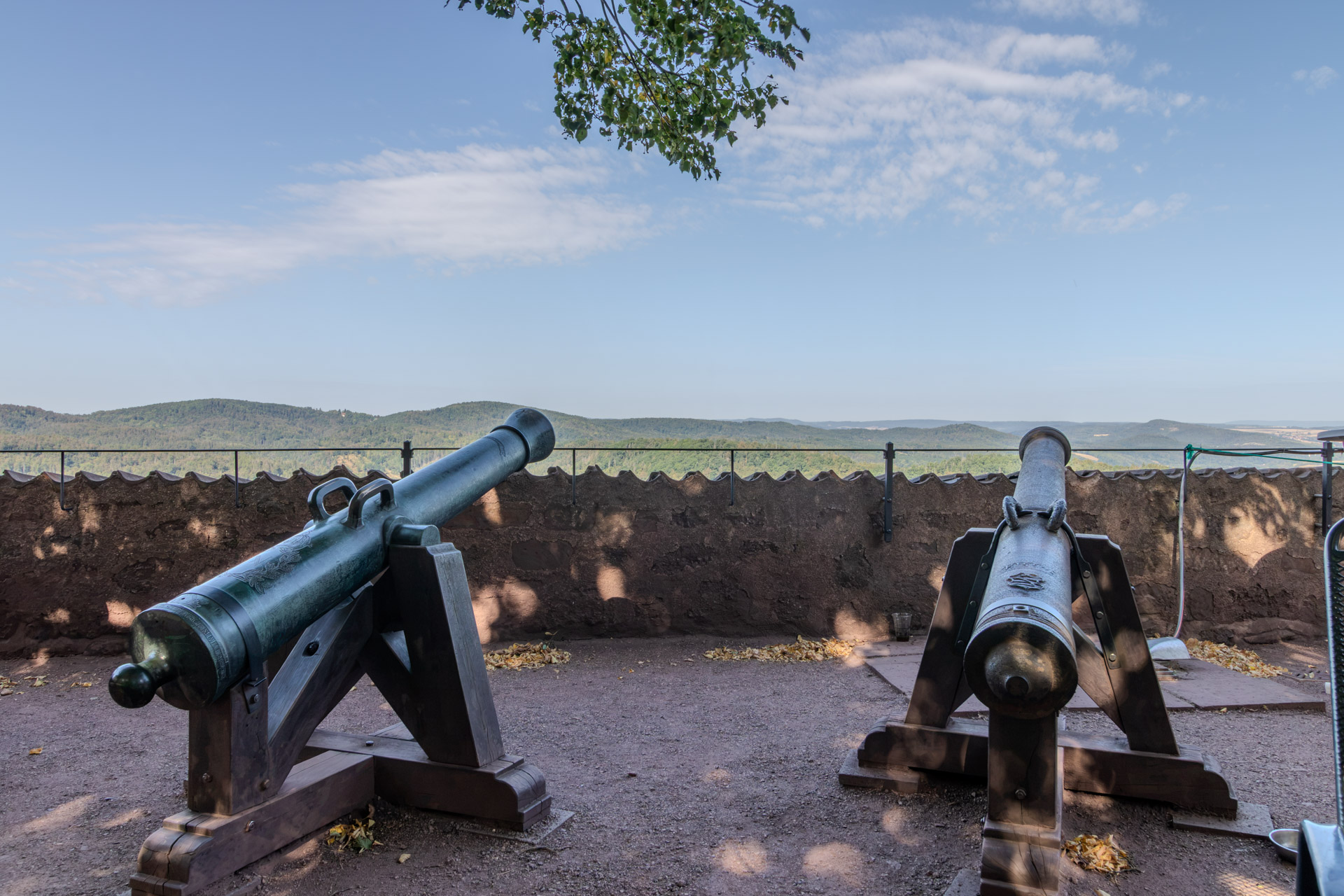Zwei alte Kanonen auf hölzernen Lafetten stehen auf einem Platz hinter einer rötlichen Burgmauer, die linke mit grüner Patina. Dahinter der Blick über bewaldete Hügel bis zum Horizont, oben ragen Zweige eines Baums ins Bild.