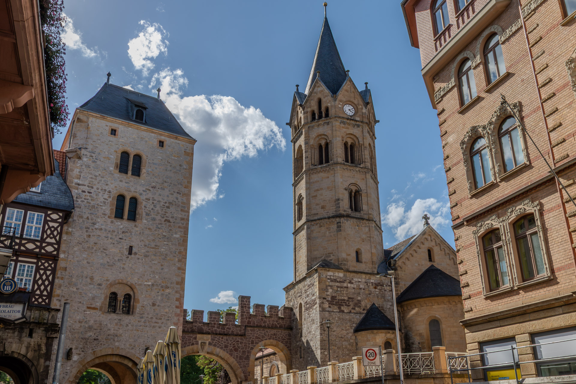 Das Nikolaitor in Eisenach, ein wuchtiger Torturm aus Bruchstein mit Schieferdach, daneben ein Torbogen mit Zinnen. Rechts dahinter der Turm der Nikolaikirche mit Uhr und spitzem Helm, am rechten Bildrand ein verziertes Backsteinhaus, links ein Fachwerkhaus. Blauer Himmel mit weissen Wolken.