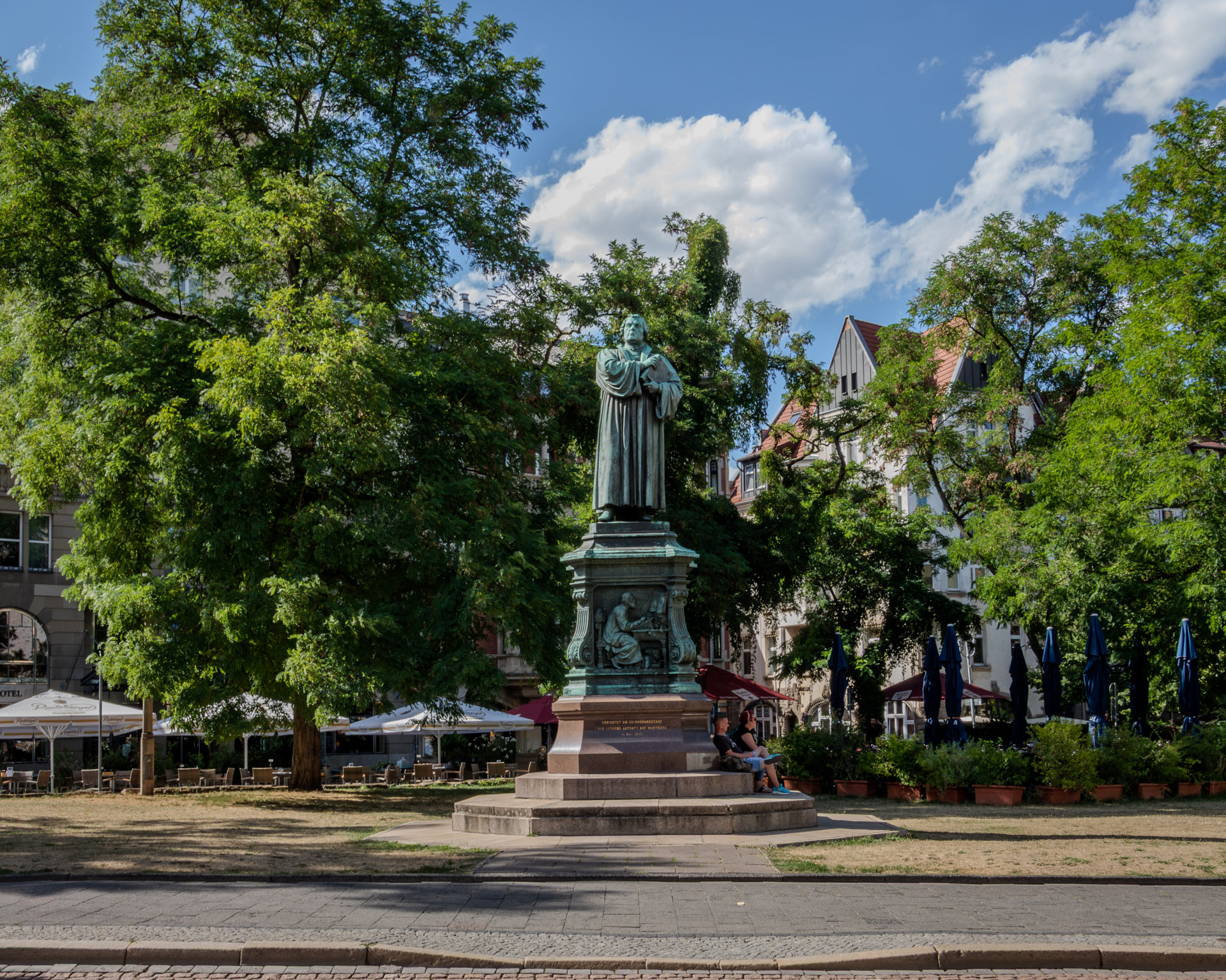 Bronzestatue von Martin Luther mit einem Buch in der Hand auf einem hohen Sockel, im Sockel ein Relief, das Luther beim Schreiben zeigt. Zwei Personen sitzen am Fuss des Denkmals, rundherum grosse Bäume, Restaurantterrassen mit Sonnenschirmen und Altbauten unter blauem Himmel mit Wolken.