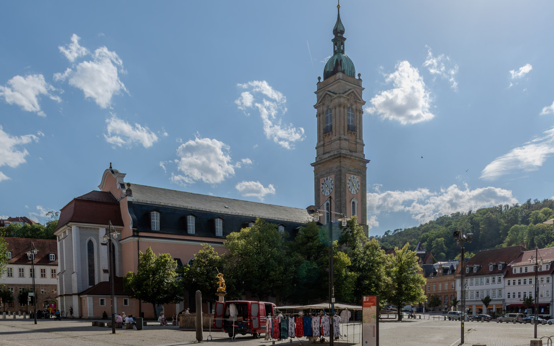 Die Georgenkirche mit rosafarbener Fassade, Schieferdach und hohem Turm mit grüner Haube und zwei Uhren, davor Bäume und ein Brunnen mit goldener Figur. Auf dem weiten Platz ein roter Lieferwagen und ein Marktstand mit Kleidern auf Ständern, rechts Häuserzeilen, dahinter ein bewaldeter Hügel.