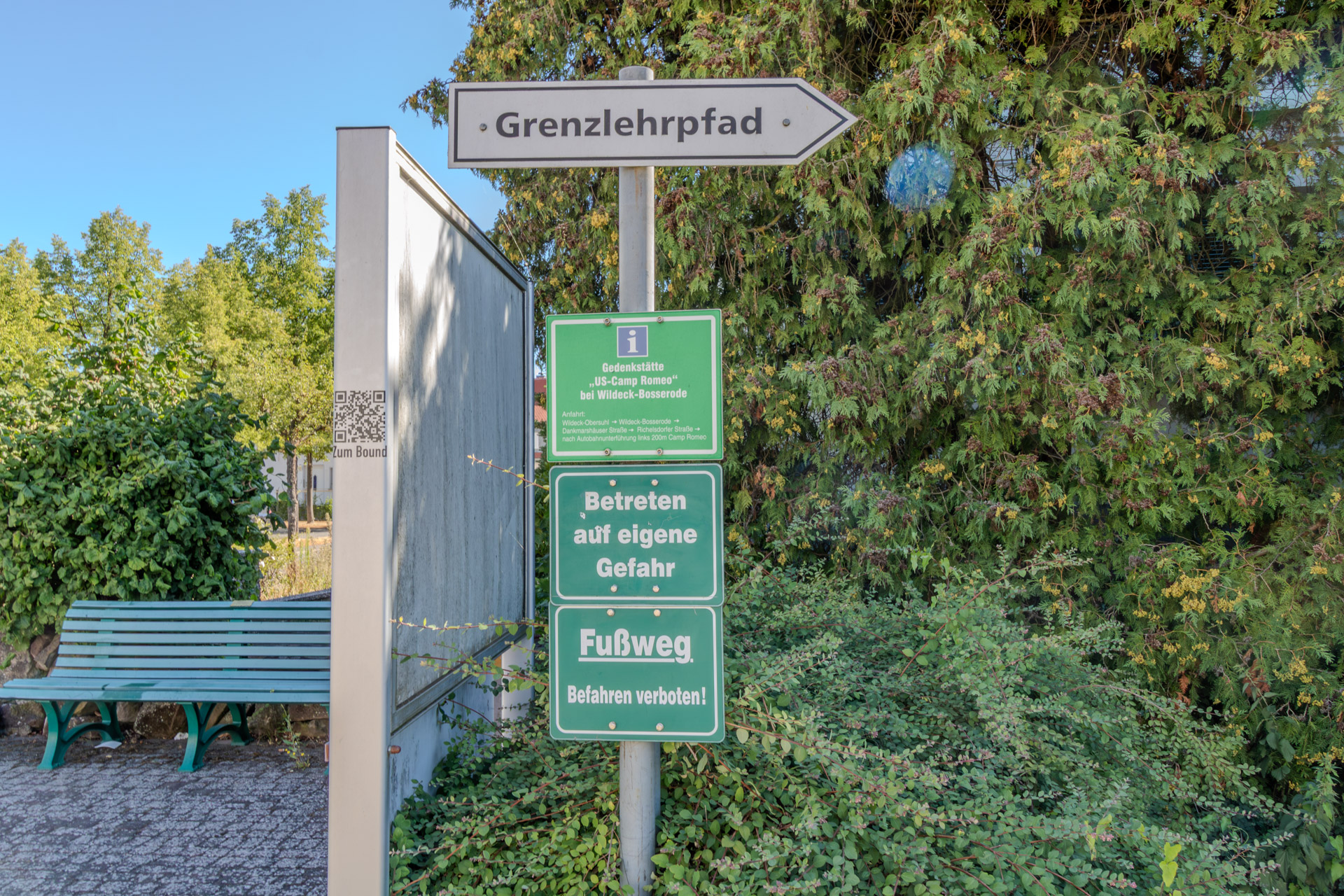 Weisser Wegweiser mit der Aufschrift «Grenzlehrpfad», darunter grüne Schilder mit einem Hinweis zur Gedenkstätte «US-Camp Romeo» bei Wildeck-Bosserode sowie «Betreten auf eigene Gefahr» und «Fussweg – Befahren verboten!». Links eine grüne Parkbank und eine Infotafel, rechts dichte Hecken.
