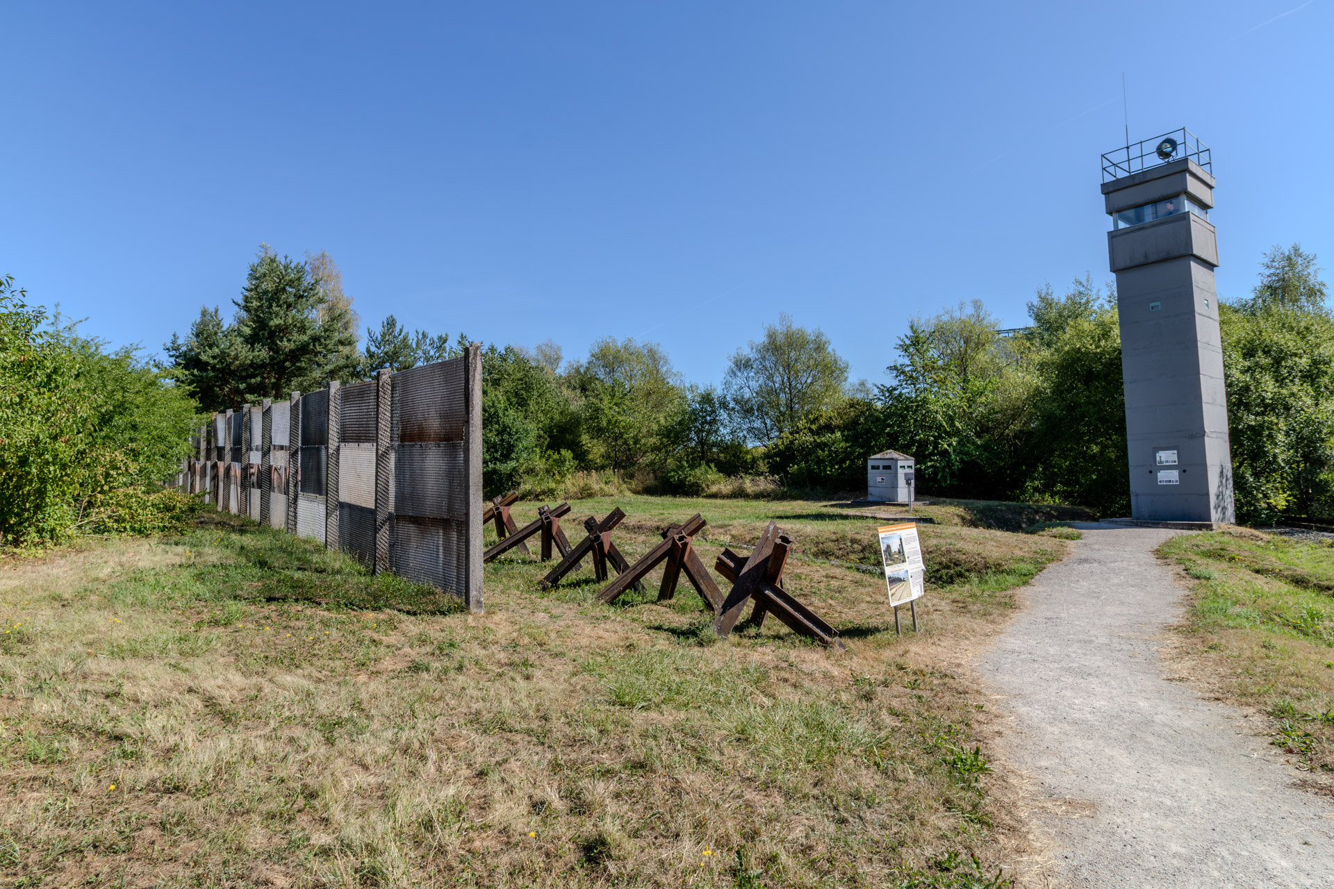 Ein Stück des ehemaligen Grenzzauns aus Metallgitter auf Betonpfosten, davor eine Reihe rostiger Panzersperren aus Stahlträgern und eine Infotafel. Rechts führt ein Weg zu einem grauen Beobachtungsturm aus Beton mit Scheinwerfer auf dem Dach, in der Mitte ein kleines Betonhäuschen, rundherum Wiese und Bäume unter wolkenlosem Himmel.