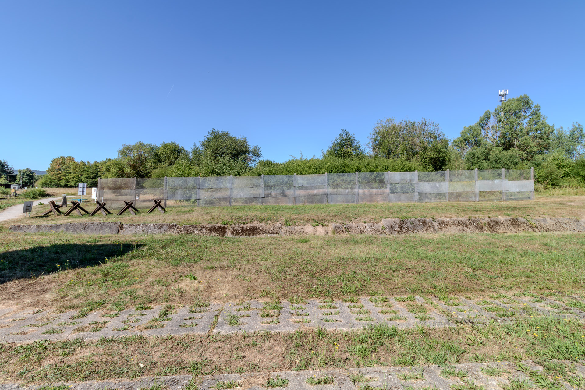 Ein langer Abschnitt des ehemaligen Grenzzauns aus Metallgitter auf einer Wiese, links davon rostige Panzersperren und Infotafeln. Davor ein mit Betonplatten befestigter Graben, im Vordergrund ein Weg aus Lochbetonplatten, zwischen denen Gras wächst. Dahinter Büsche und Bäume unter wolkenlosem blauem Himmel.
