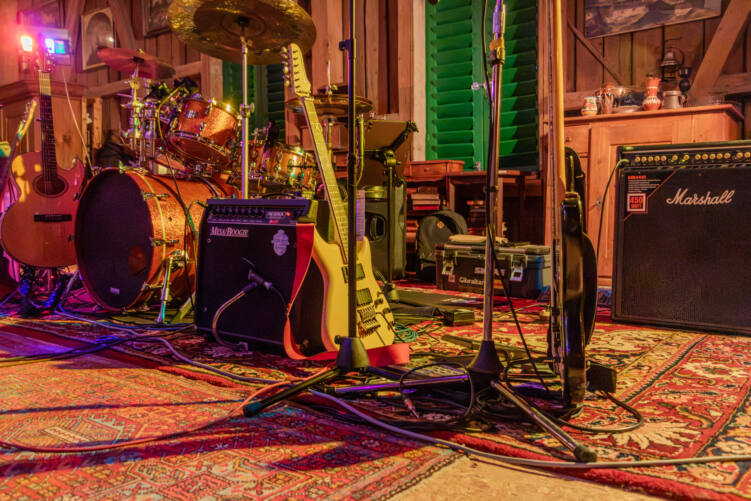 Instrumente auf Orientteppichen in der Antikscheune, aufgenommen auf Bodenhöhe. In der Mitte lehnt eine gelbe E-Gitarre mit rotem Gurt an einem Mesa/Boogie-Verstärker, dahinter ein kupferfarbenes Schlagzeug, links eine Akustikgitarre, rechts ein schwarzer Bass vor einem Marshall-Verstärker, dazwischen Mikrofonständer und Kabel. Im Hintergrund Holzwände, grüne Fensterläden und ein alter Schrank mit Krügen und Vasen, oben links ein violetter Scheinwerfer, alles im warmen Licht.